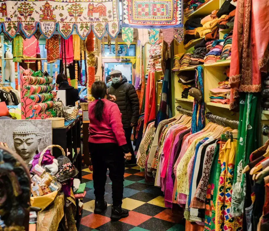 A colorful and bustling clothing store in Vijaynagar, Mysore. The photo shows a person in a pink sweater standing in the aisle, surrounded by racks of clothes and shelves of various items. This image could be used by Aspire Digital Solutions to demonstrate their expertise in digital marketing for local retail businesses.