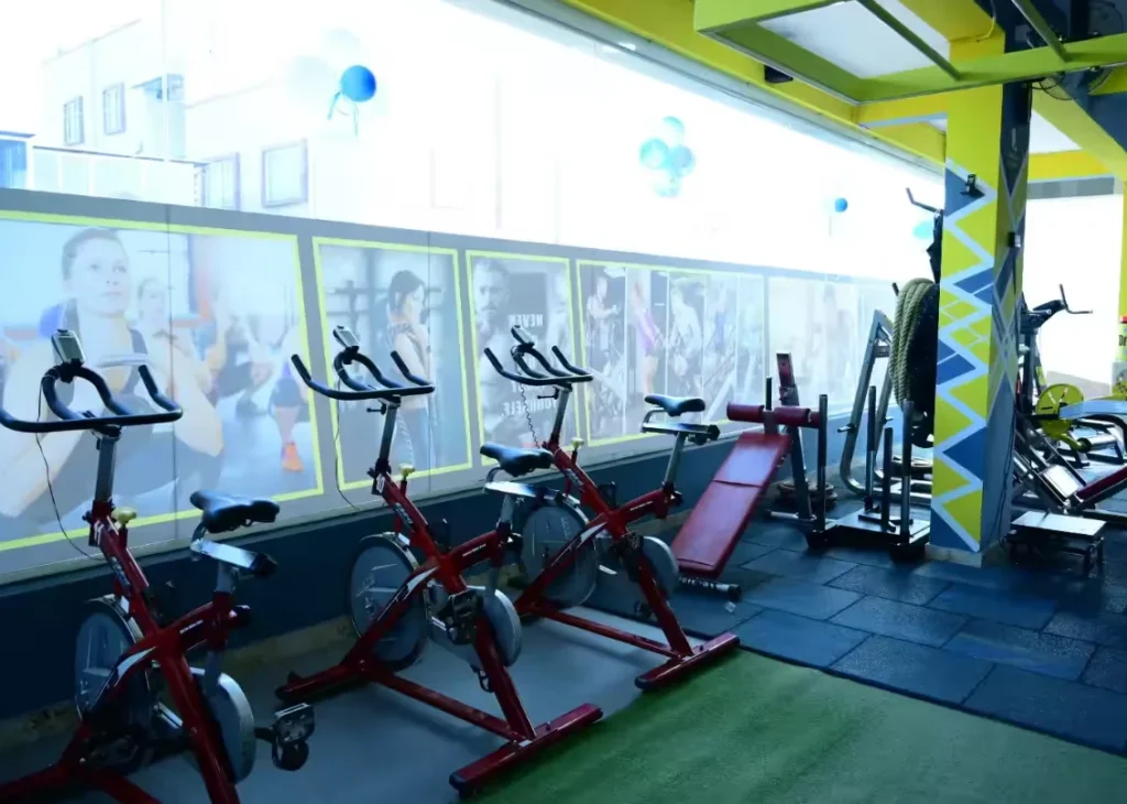 A row of spin bikes and other fitness equipment stand ready for use inside a gym. The walls are decorated with large, backlit posters of people working out. The image could be used to promote a fitness center in J.P. Nagar, Mysore, or to showcase the digital marketing work of a company like Aspire Digital Solutions for clients in the fitness industry.